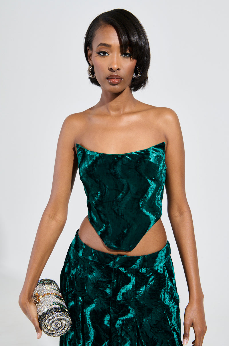 A person with short black hair and statement earrings stands against a plain background, elegantly dressed in the GREEN WITH ENVY VELVET CORSET TOP—a strapless two-piece crushed velvet outfit featuring a corset top with a pointed hem that seamlessly matches the skirt, while holding a beaded clutch. 1
