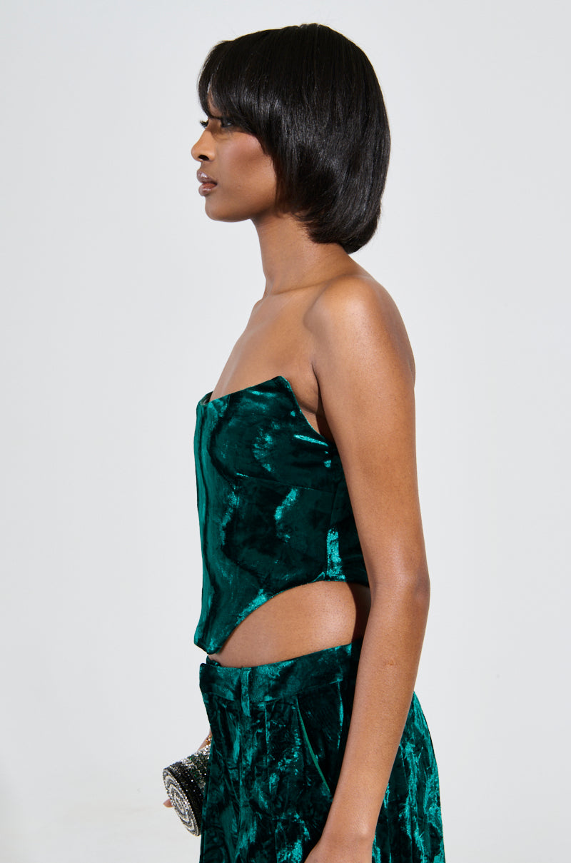 A person with shoulder-length hair is sporting the GREEN WITH ENVY VELVET CORSET TOP in dark green crushed velvet, complemented by matching pants. They hold a small, embellished clutch in one hand against a plain, light-colored background. 2