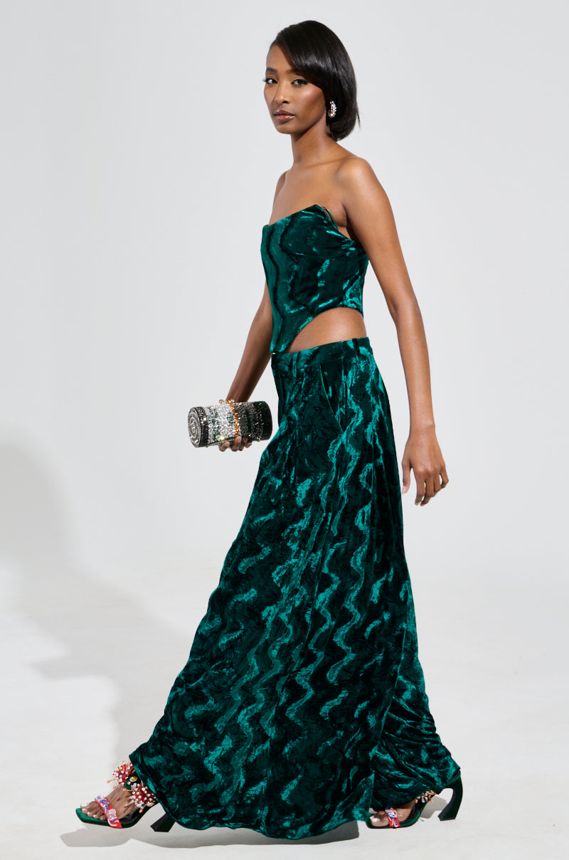 A person in the GREEN WITH ENVY VELVET CORSET TOP, featuring an elegant strapless silhouette with a side cutout, holds a glittering clutch. They wear decorative heels and have long, dark hair, standing confidently against a plain background. 4