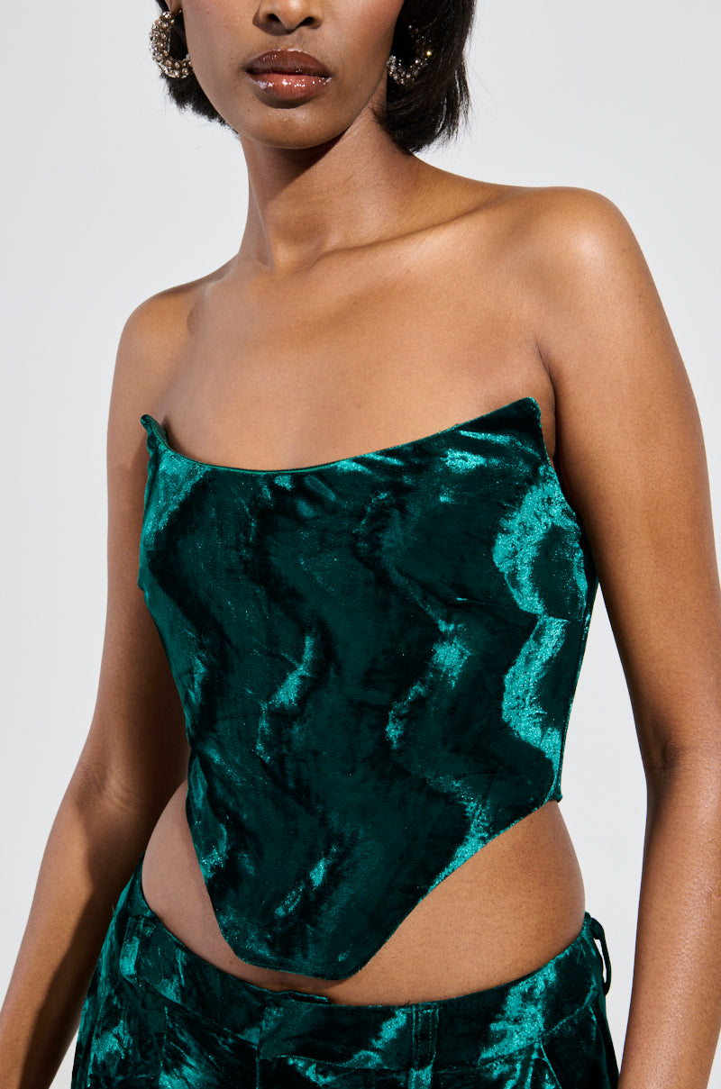 A woman is wearing the Green With Envy Velvet Corset Top and matching pants. The crushed velvet catches the light beautifully, and her hair falls in loose waves as she stands against a neutral background, exuding elegance with just a hint of mystery in her striking silhouette. 7