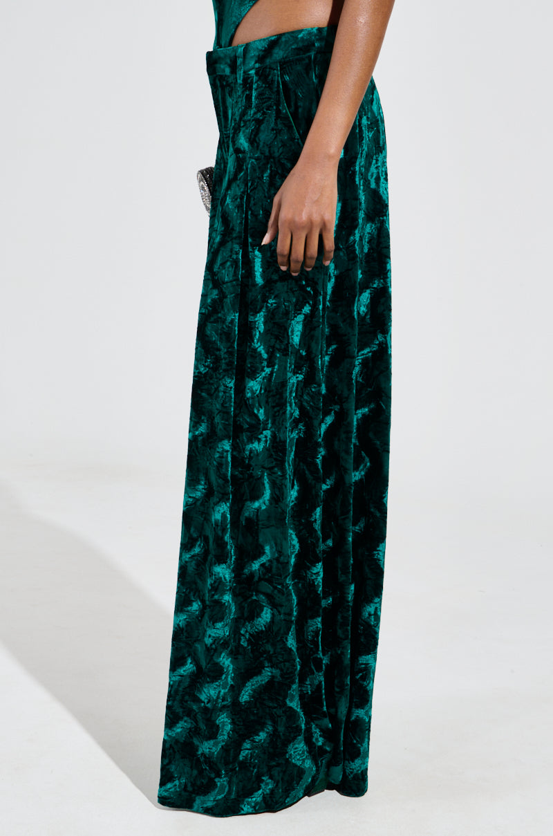 A person wearing a pair of "Green With Envy" velvet wide leg trousers, featuring a textured crushed velvet fabrication and a high-rise waist. The person's left hand rests gently on the hip against the plain, light background. 3