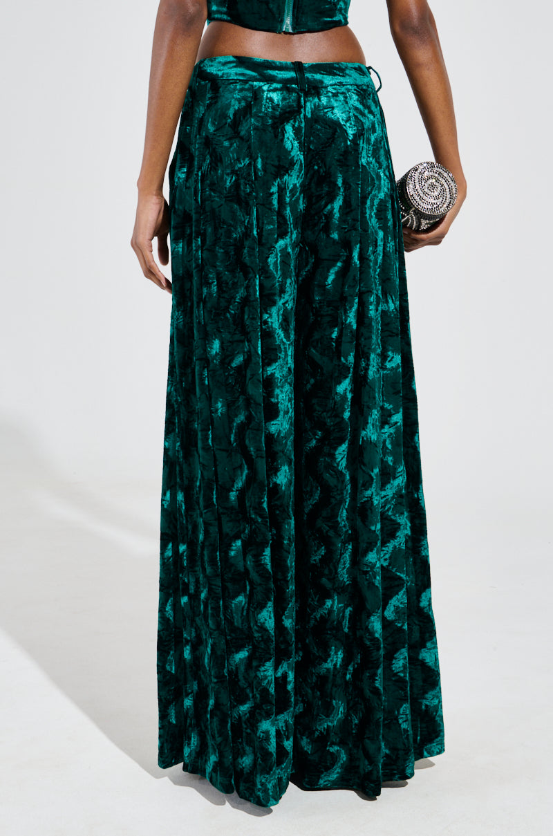 A person wearing GREEN WITH ENVY velvet wide-leg trousers stands against a plain background, holding a small, round embellished clutch in their left hand. 4