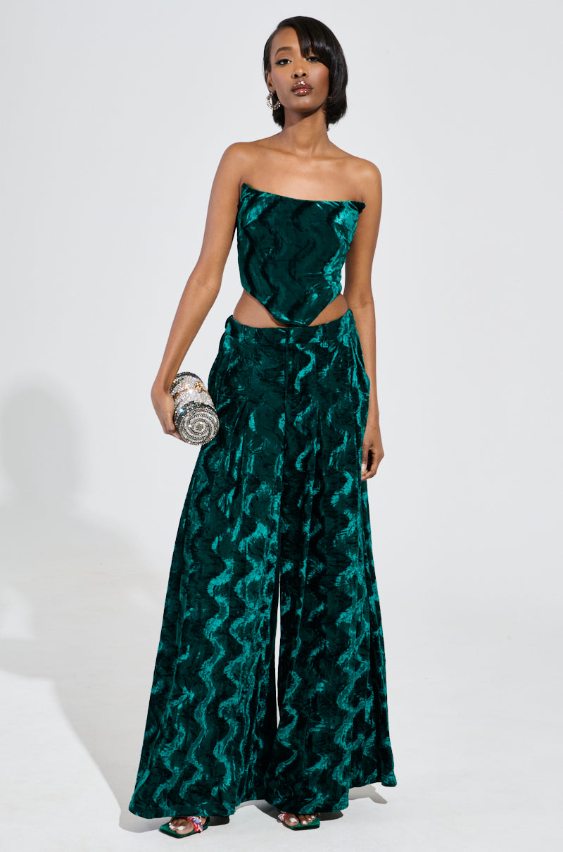 Wearing the GREEN WITH ENVY VELVET WIDE LEG TROUSER, a woman poses against a plain white background. The ensemble is strapless and features an eye-catching emerald green color with crushed velvet fabrication, complemented by cut-out details at the high-rise waist and wide-leg pants. She accessorizes with a sparkly clutch. 5
