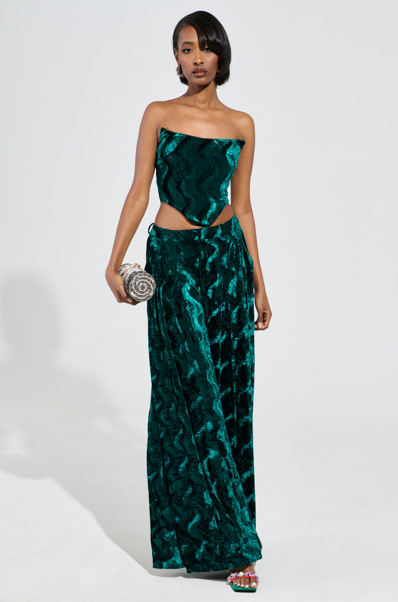 A woman stands in a chic ensemble, wearing the GREEN WITH ENVY VELVET WIDE LEG TROUSERS that feature a high-rise waist and a strapless top. She accessorizes with a patterned clutch and colorful sandals, all set against a plain white background. 6