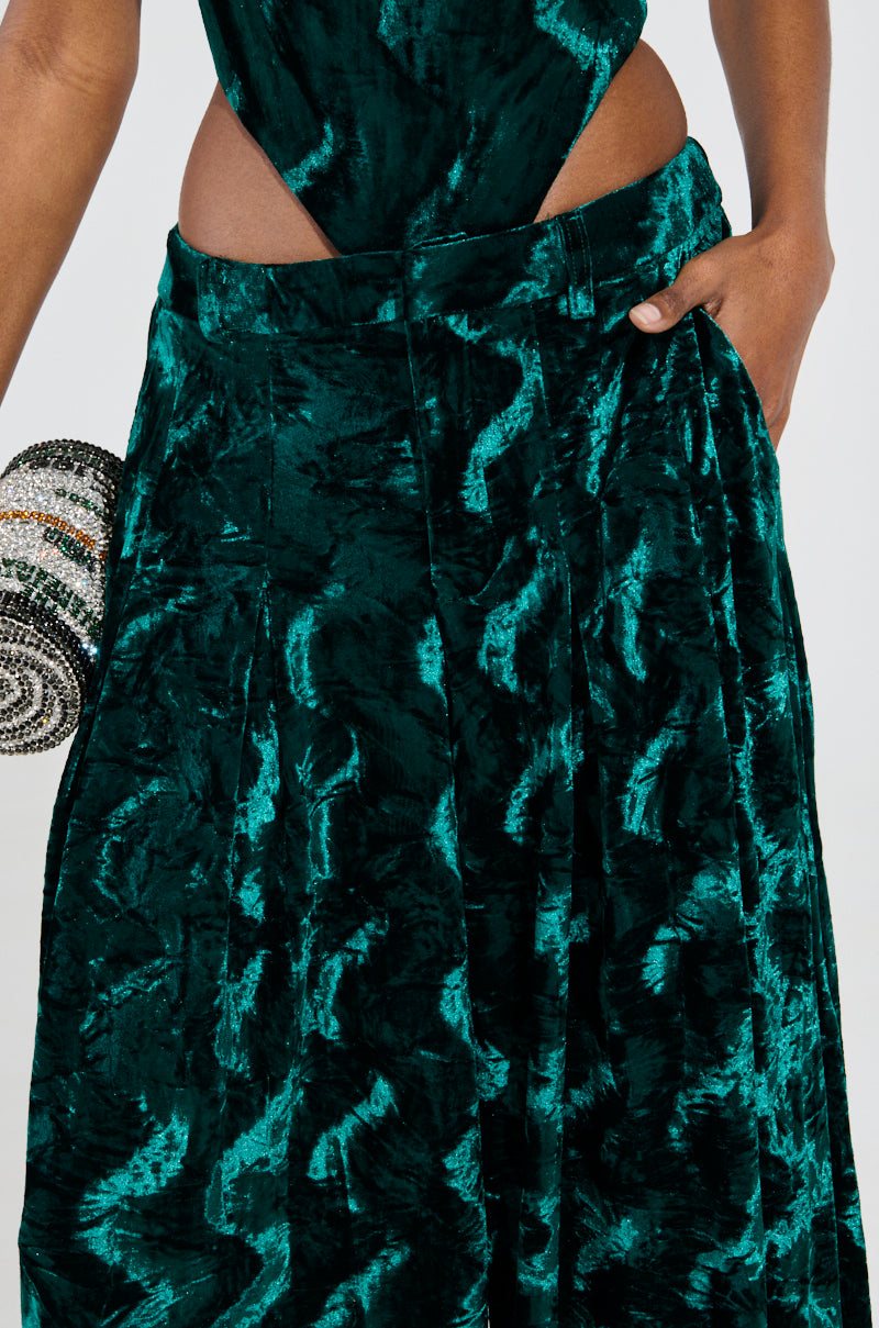 A person in emerald green velvet wide leg trousers with a side cutout, accessorized with a small, round, beaded clutch. The crushed velvet fabric adds depth with textured patterns, and their hand near a pocket exudes casual elegance. 7