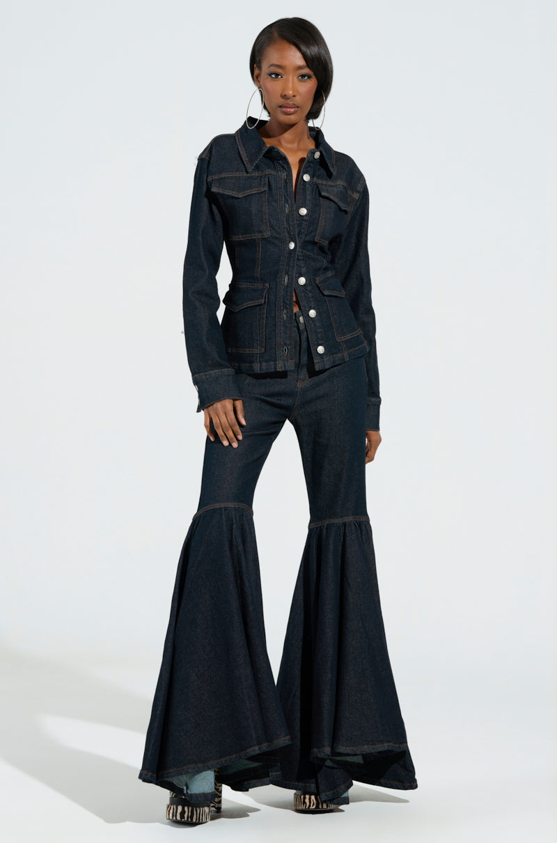 A person confidently stands in a dark wash denim outfit, featuring the EVERYDAY LIVIN' DENIM BUTTON DOWN SHIRT with large pockets and flared bell-bottom jeans—a perfect vibe for Everyday Livin'. The light, neutral background highlights their style, complemented by large hoop earrings and patterned shoes.