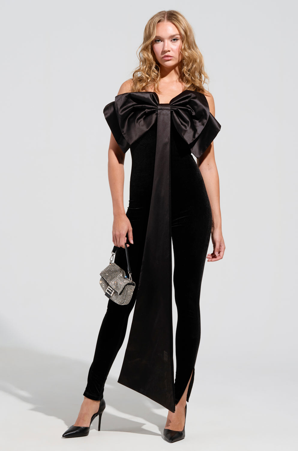 Clad in the stylish "I'M THE GIFT" velvet jumpsuit with adjustable straps and an eye-catching satin bow, a woman stands confidently. She accessorizes with a small silver handbag and black high heels. Her long, wavy hair cascades freely as she poses against a minimalist light background.