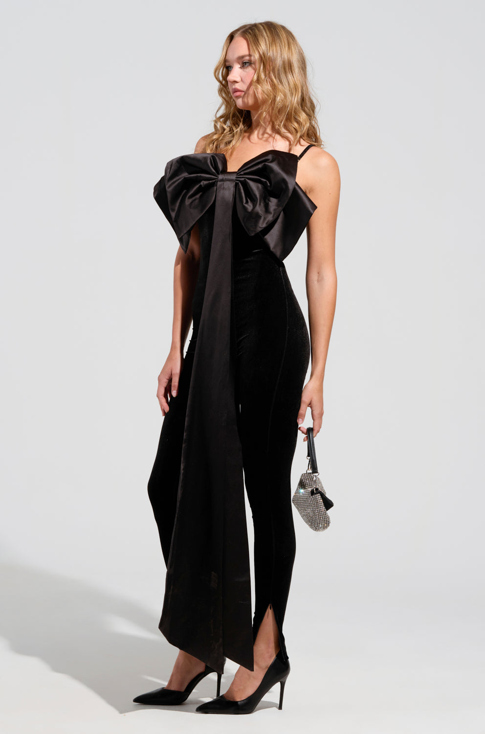 A woman in the I'M THE GIFT velvet jumpsuit, featuring adjustable straps and a prominent satin bow, stands against a light background. She is accessorized with a small silver handbag and wears black high heels. Her hair is styled in loose waves, completing the elegant look.