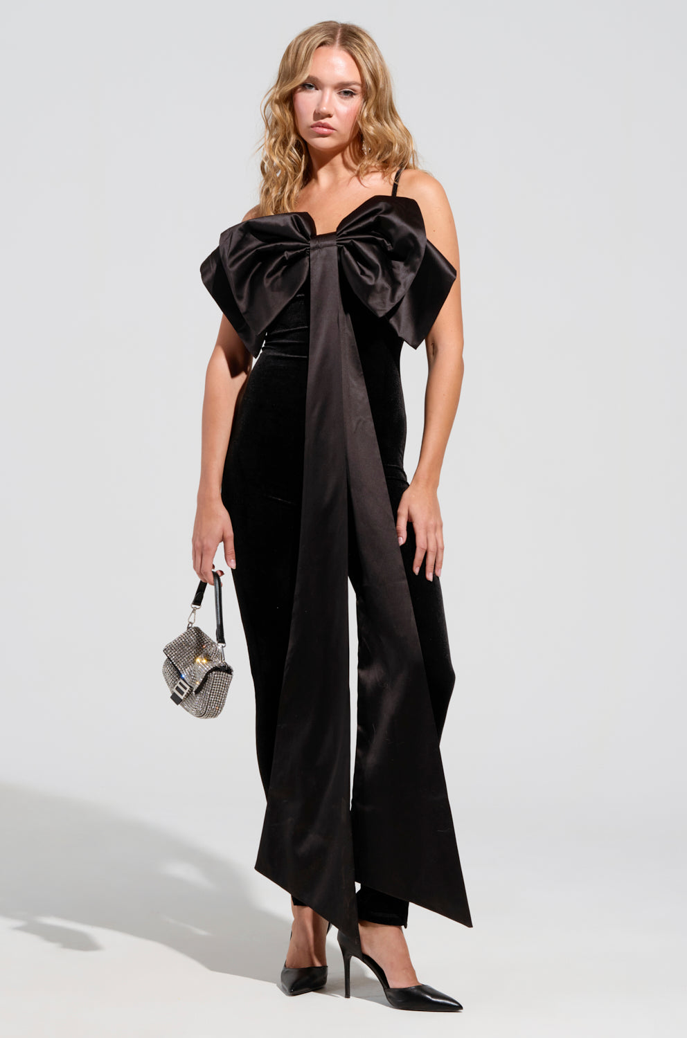 A woman in the I'M THE GIFT Velvet Jumpsuit with a Bow stands against a plain background. She holds a small handbag and wears pointed black heels, while her blonde hair is styled in loose waves.