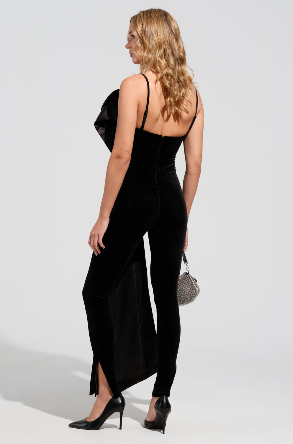 A woman with wavy blonde hair stands in profile, emphasizing her I'M THE GIFT velvet jumpsuit with adjustable straps and high heels. She carries a small, round silver handbag against a plain, light-gray background.