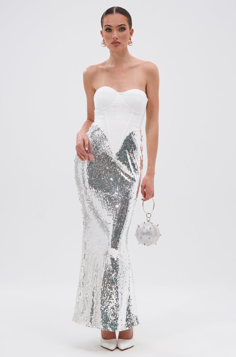 A woman poses against a plain white background wearing the TOO GOOD FOR THIS MAXI DRESS IN WHITE, featuring a strapless corset-style bodice and a shiny silver sequin maxi skirt, styled with a round silver handbag and white heels.