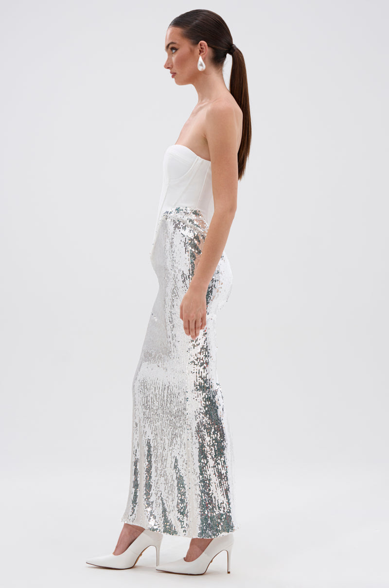 A woman stands in profile against a white background wearing the TOO GOOD FOR THIS MAXI DRESS IN WHITE, featuring a strapless corset-style bodice and silver sequin maxi skirt, styled with white heels and large earrings. Her hair is in a sleek ponytail.