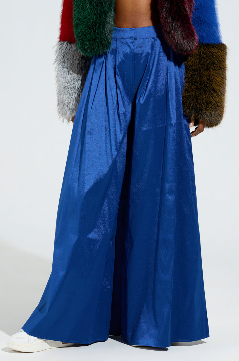 A person wearing the SHALLOW TROUSER in jewel-tone, wide-leg satin stands against a light background. They are also sporting a colorful, textured jacket featuring a patchwork of red, green, and blue sections paired with white sneakers.