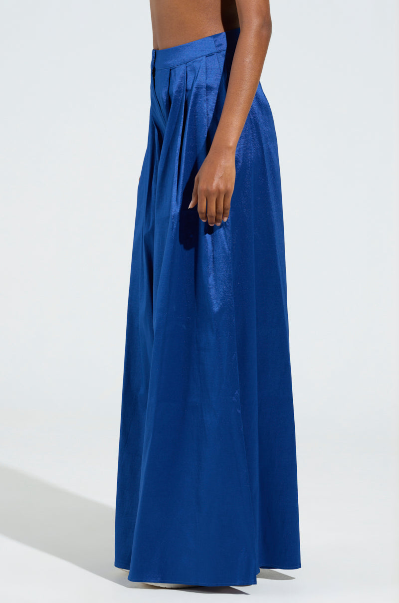 A person is wearing SHALLOW TROUSER, a high-waisted, blue full-length skirt with pleats, standing against a plain background. The jewel-toned bottom appears flowy and elegant, complementing the person's silhouette.