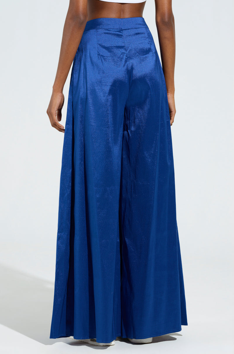 A person wearing the SHALLOW TROUSER high-waisted, jewel-toned wide-leg satin pants in vibrant blue stands against a light background. The image focuses on the lower half, highlighting the flowing silhouette and shimmering sheen of the fabric.