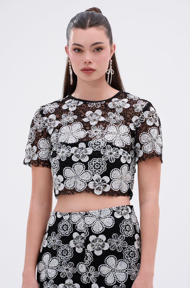 A woman with long, dark hair in a high ponytail wears the FLORAL SEQUIN MESH SHORT SLEEVE BLOUSE, featuring striking white floral embroidery, and poses against a plain light background.