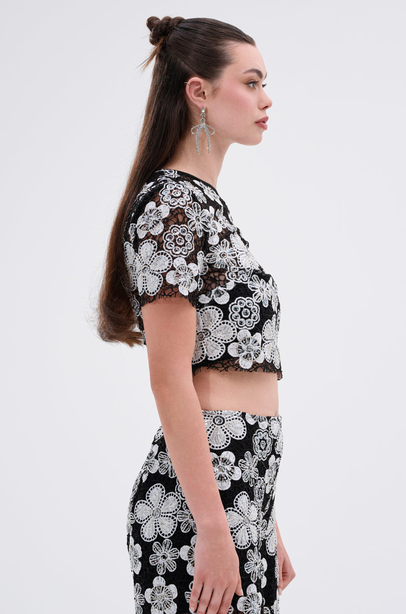 A woman with long brown hair in a half-up style models the FLORAL SEQUIN MESH SHORT SLEEVE BLOUSE, paired with statement earrings, standing in profile against a plain white background.