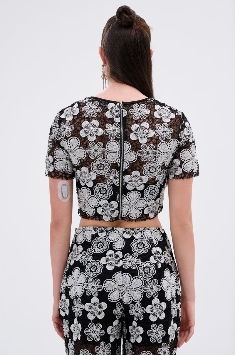 A person with long brown hair, seen from behind, wears the FLORAL SEQUIN MESH SHORT SLEEVE BLOUSE in black with white floral detailing; a medical device is visible on their upper left arm.