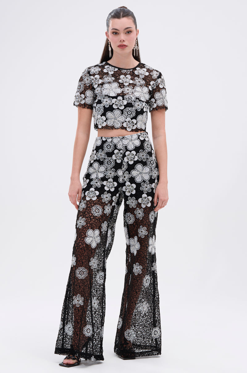 A woman faces forward in the FLORAL SEQUIN MESH SHORT SLEEVE BLOUSE and matching high-waisted pants with bold white floral embroidery, black sandals, hair pulled back, against a plain white background.