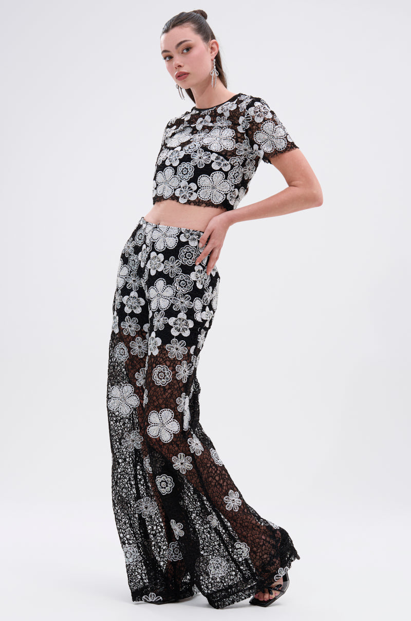 A woman poses against a white background wearing the FLORAL SEQUIN MESH SHORT SLEEVE BLOUSE with matching wide-leg pants featuring striking white floral embroidery. Her hair is pulled back, and she accessorizes with earrings and sandals.
