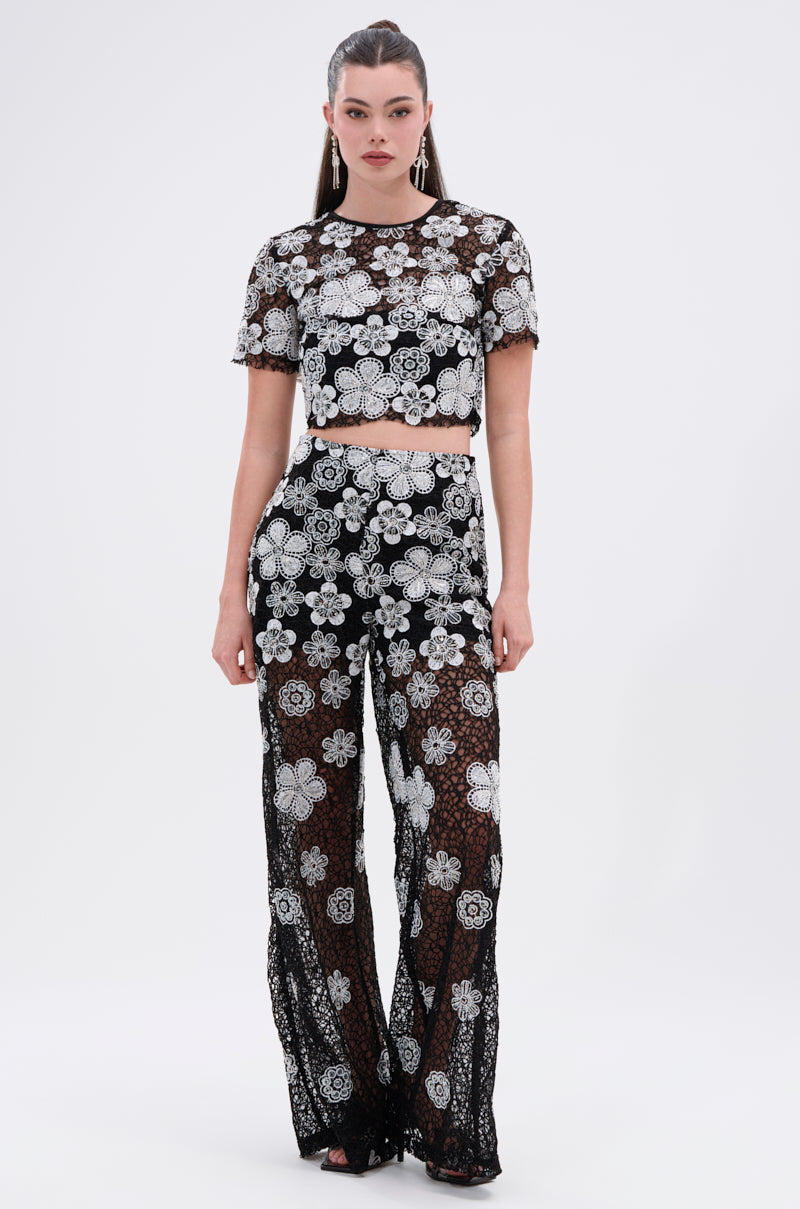 A woman poses confidently against a plain white background, wearing the FLORAL SEQUIN MESH SHORT SLEEVE BLOUSE paired with high-waisted pants. Her long hair is tied back, and the sheer blouse features intricate white floral details.