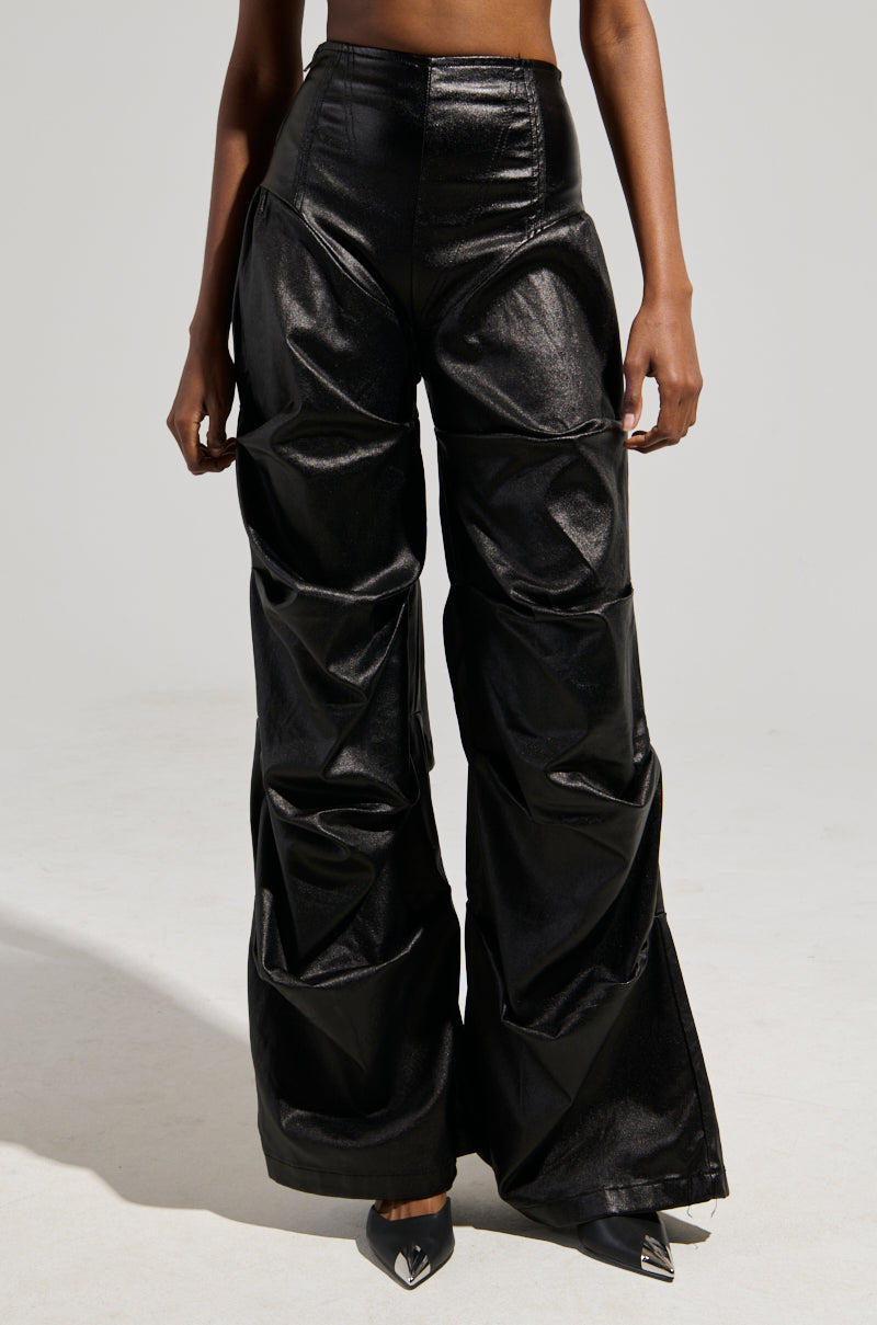 A person is wearing the MIXED MESSAGES FAUX LEATHER PANT in black, featuring a shiny, high-waisted wide-leg design with a textured ruched pattern, embodying a sophisticated blend of style and comfort. Their pointed-toe black shoes complement the look against the plain, neutral backdrop.