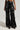 A person is wearing the MIXED MESSAGES FAUX LEATHER PANT in black, featuring a shiny, high-waisted wide-leg design with a textured ruched pattern, embodying a sophisticated blend of style and comfort. Their pointed-toe black shoes complement the look against the plain, neutral backdrop.