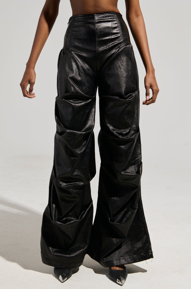 A person wearing the MIXED MESSAGES FAUX LEATHER PANT, characterized by its black, shiny faux leather high-waisted wide-leg design with a ruched detail, is standing against a plain gray background, showcasing only their torso and legs.