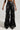 A person wearing the MIXED MESSAGES FAUX LEATHER PANT, characterized by its black, shiny faux leather high-waisted wide-leg design with a ruched detail, is standing against a plain gray background, showcasing only their torso and legs.