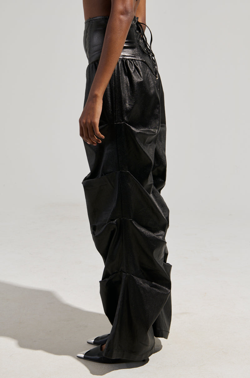 A person stands sideways wearing the MIXED MESSAGES FAUX LEATHER PANT, featuring an oversized black silhouette with lace-up details at the waist. The high-rise drop waist design results in pronounced folds, and the wide-leg flared style flows down to the floor, concealing their feet. The background is plain and light-colored.