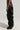 A person stands sideways wearing the MIXED MESSAGES FAUX LEATHER PANT, featuring an oversized black silhouette with lace-up details at the waist. The high-rise drop waist design results in pronounced folds, and the wide-leg flared style flows down to the floor, concealing their feet. The background is plain and light-colored.