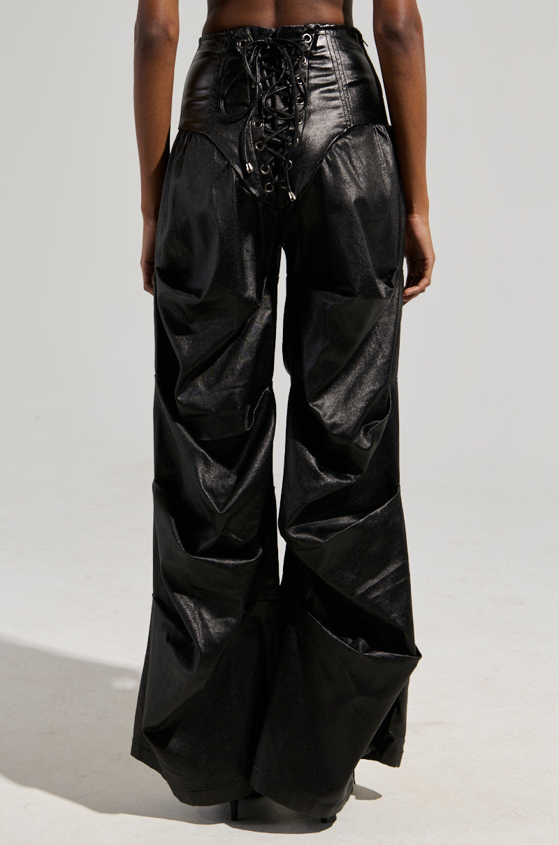 A back view of a person wearing the MIXED MESSAGES FAUX LEATHER PANT in black, showcasing its wide-legged design and shiny texture. The pants include a lace-up detail and high waist for a stylish and unique appearance. The background is plain light gray.