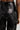 A person wearing the MIXED MESSAGES FAUX LEATHER PANT showcases a close-up of its details, highlighting the lace-up design on the back. The high-rise drop waist enhances its sleek fit, while the shiny faux leather fabric reflects light, adding a glossy texture.