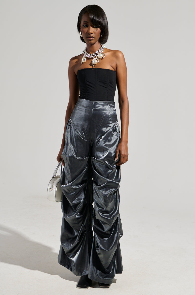 A person wearing a strapless black top, statement silver necklace, and Bella Satin High Rise Trouser in grey with a wide barrel-legged silhouette stands against a plain background, holding a small silver bag.