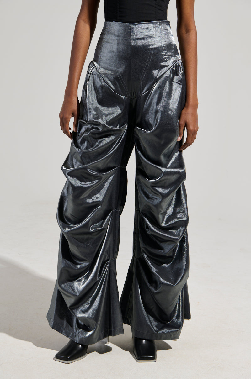 A person wearing shiny Bella Satin High Rise Trousers in grey, featuring a wide barrel-legged silhouette with gathered and layered fabric, stands against a neutral background. They complete the look with a black top and black square-toed shoes.