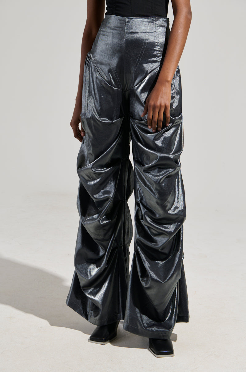 Wearing the Bella Satin High Rise Trouser in Grey, a person showcases shiny, dark metallic pants with a distinctive ruched design. These trousers feature a high-rise cut with a wide barrel-legged silhouette that elegantly complements a black top. The neutral background highlights the unique texture and fashion-forward style of the pants.