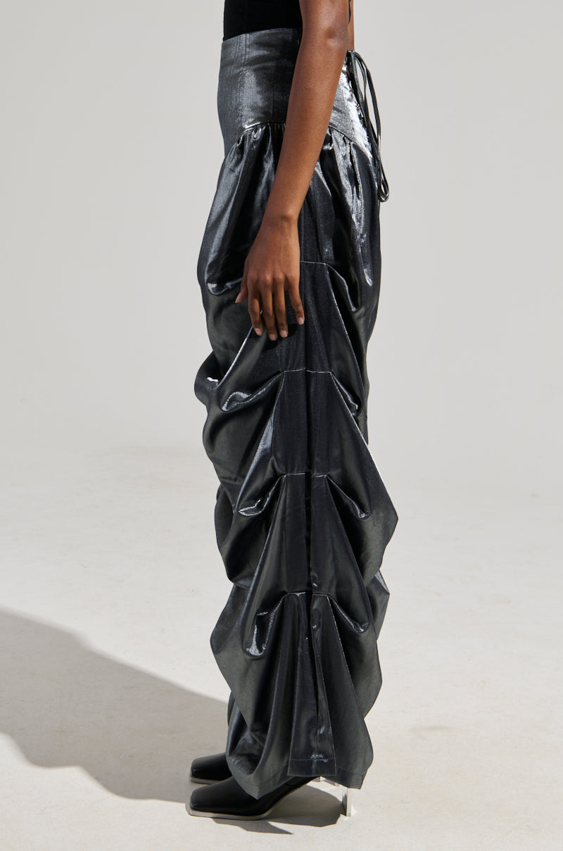 A person is wearing the high-rise Bella Satin Trousers in grey, showcasing a metallic silver sheen with shiny, ruched details. The trousers feature a distinctive wide barrel-legged silhouette that adds texture to the outfit. To complete the look against a plain, light-colored background, they pair it with black shoes as they stand sideways.