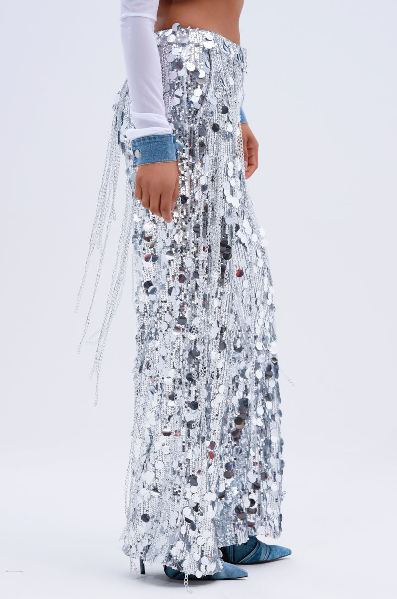 Dressed in EXPENSIVE TASTE STRETCHY SEQUIN CHAIN PANTS and a fitted white long-sleeve top, the person shines. They wear blue heeled shoes against a solid white background.