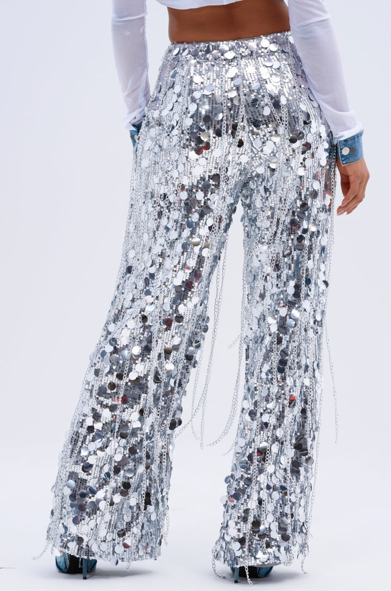 Someone is wearing the EXPENSIVE TASTE STRETCHY SEQUIN CHAIN PANTS featuring hanging chains, paired with a white long-sleeve top. The shimmering pants contrast beautifully against the plain light background.