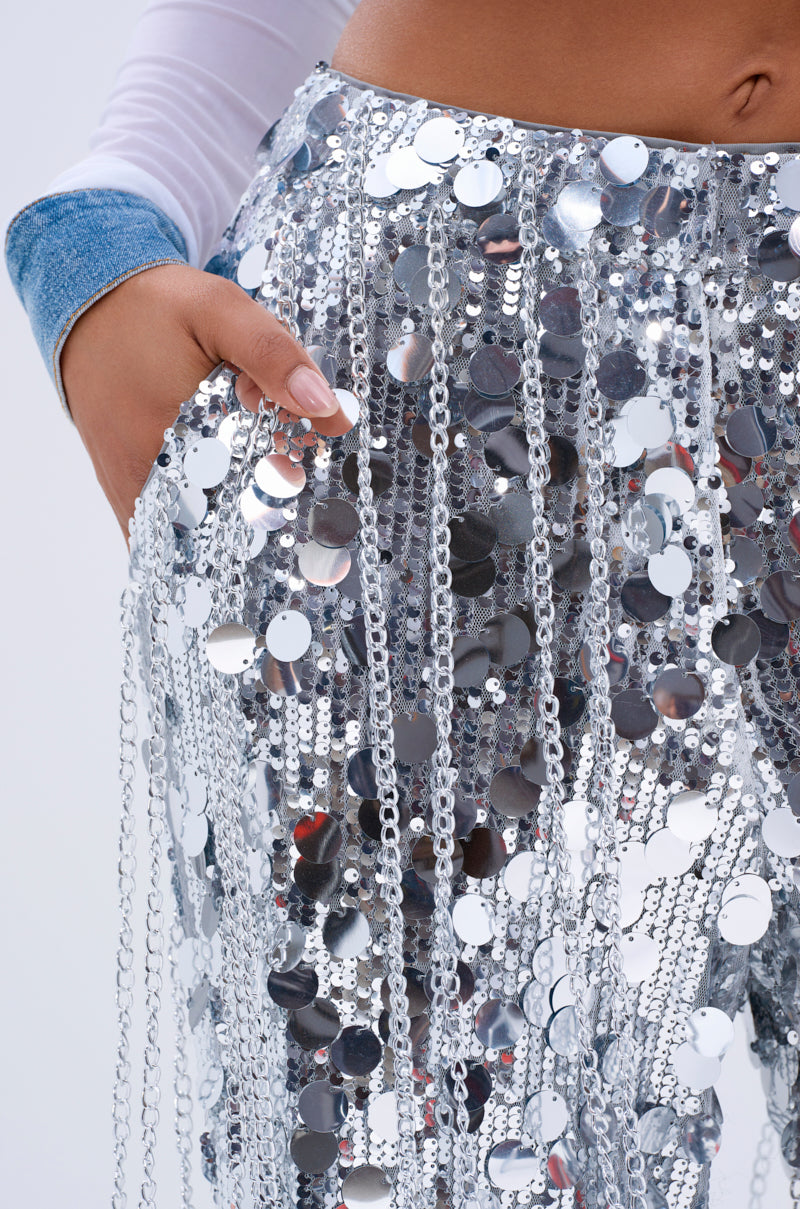 Wearing the EXPENSIVE TASTE STRETCHY SEQUIN CHAIN PANTS, a person adorned in high-rise sequin pants with silver fringes and chain link details holds their waist. The sequins shimmer beautifully, while a hint of a long-sleeved top peeks through.