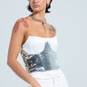 A person with short dark hair wears a white strapless dress featuring a sparkling silver sequin bodice. They have shoulder tattoos and accessorize with a chain necklace and the FOR THE NIGHT SEQUIN UNDERBUST BELT. The background is plain and light-colored.