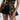 Person wears a camouflage corset with a lace front over black shorts and top, holding a strap, accessorized with the SEE THRU YOU CAMO PEPLUM BELT. The stylish, military-inspired outfit is cropped to show the torso and part of the arm against a light gray background.