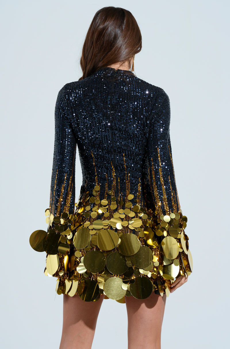 A woman with long brown hair is wearing the "Make a Statement" Sequin Mini Dress, featuring a curve-skimming silhouette adorned with large gold sequins. Her back faces the camera against a light blue background.