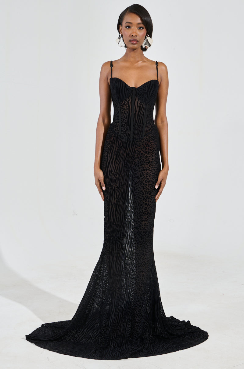 A person wearing the "ON YOUR FYP" printed velvet burnout maxi dress strikes a pose against a plain background. The long, black gown showcases sleeveless spaghetti straps, sheer mesh fabric, a fitted bodice, and a flowing train with a delicate animal print design. They have short hair and accessorize with hoop earrings.