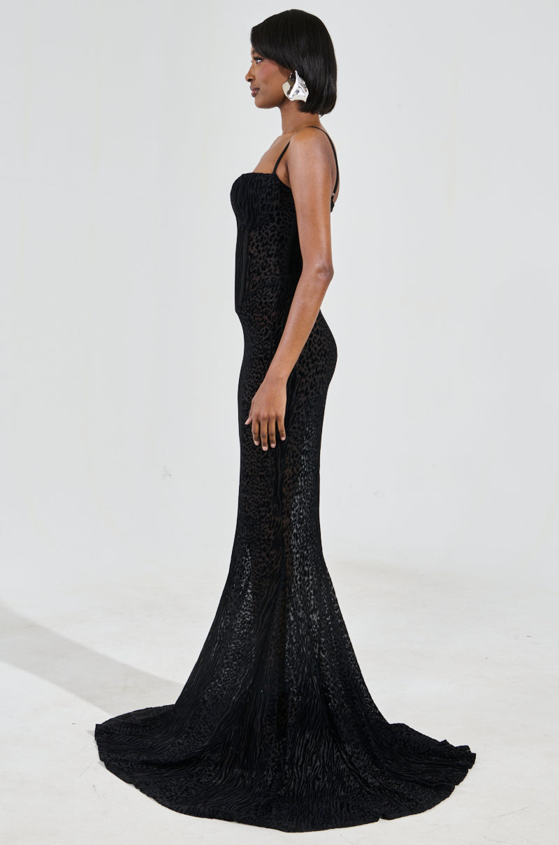 A woman stands in profile, showcasing the ON YOUR FYP PRINTED VELVET BURNOUT MAXI DRESS, a fitted, strapless black evening gown adorned with a textured animal print design. Her short hair is styled with a white accessory, and the gown boasts a long, flowing train. The plain, light-colored background accentuates the elegance of the dress.