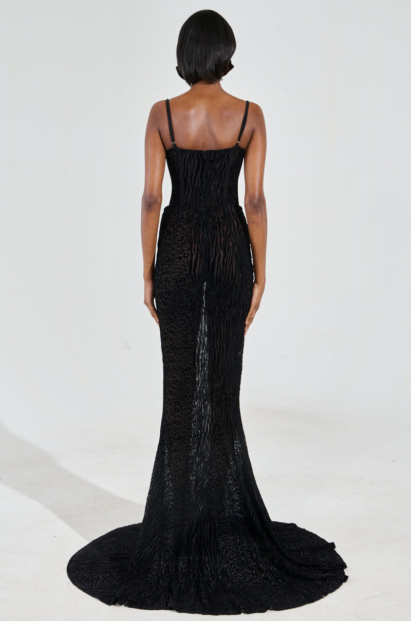 A woman stands with her back turned, wearing the ON YOUR FYP PRINTED VELVET BURNOUT MAXI DRESS. This elegant black maxi dress features thin straps, a subtle animal print design, and a train that trails along the floor. Her hair is styled in a sleek bob against a plain light gray background.