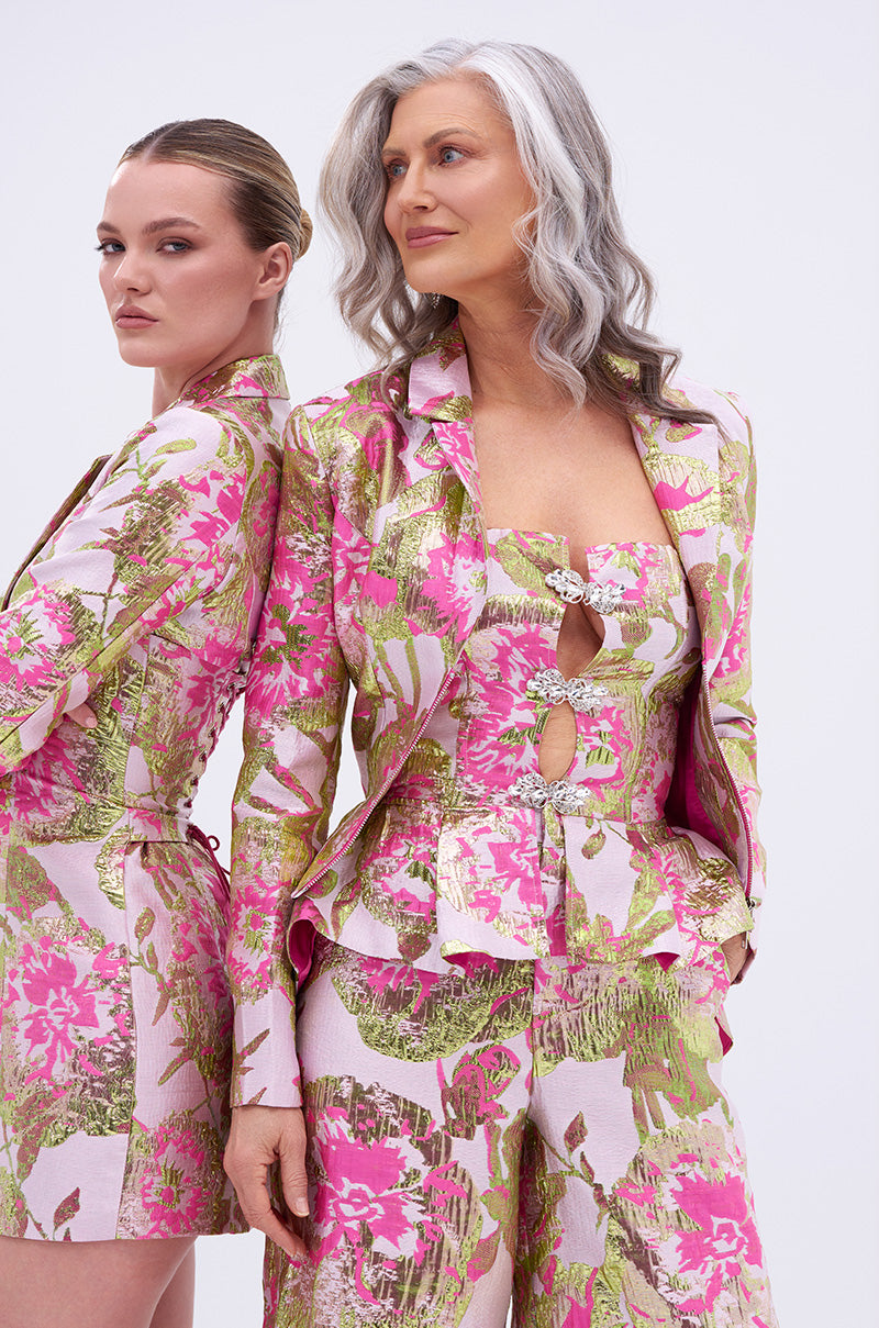 Two women stand back-to-back in vibrant pink, green, and gold LOVELY FLORAL BROCADE BLOUSE styles. One has long gray hair; the other wears a low bun. Both look confidently off-camera against a plain white background.