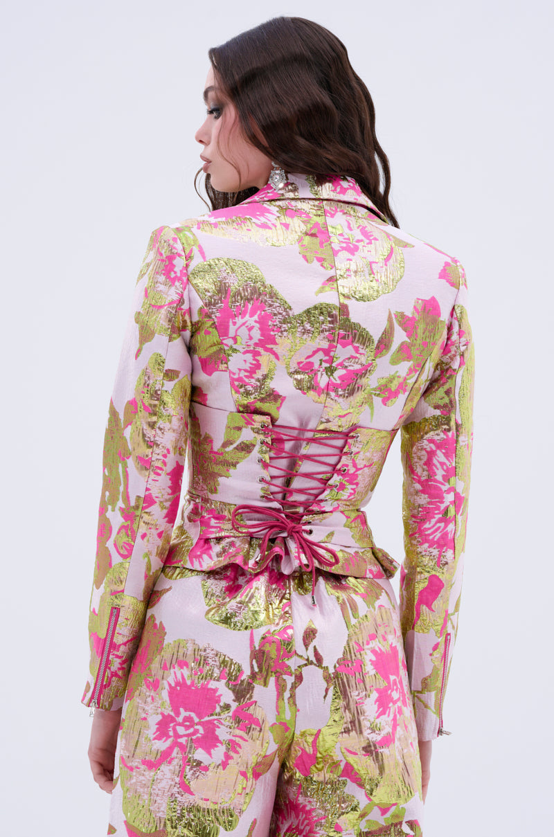 A woman is seen from the back wearing the LOVELY FLORAL BROCADE BLOUSE in pink, green, and gold, featuring corset-style lacing, a waist bow detail, and a luxurious satin-lined interior.