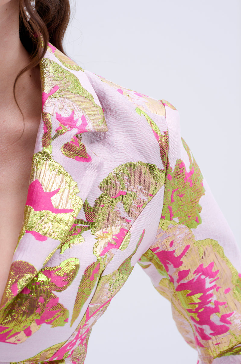 A close-up of a woman in the LOVELY FLORAL BROCADE BLOUSE, featuring light pink fabric with shiny gold and bright pink floral patterns. The image highlights the lapel, shoulder, and upper chest area.
