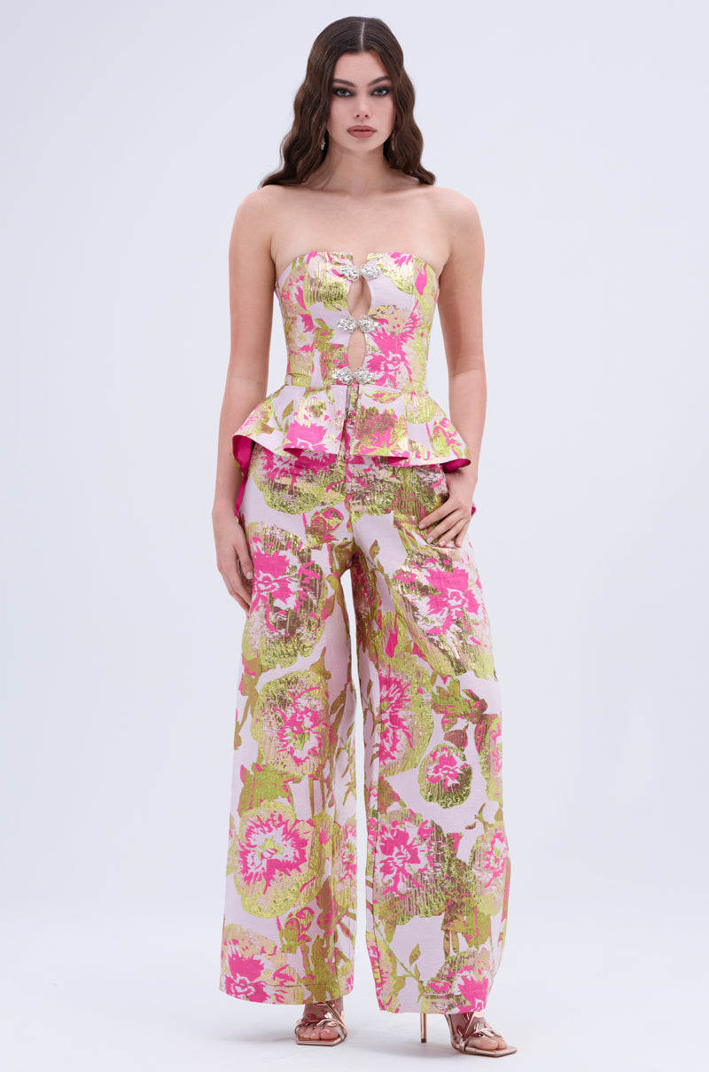 A woman is wearing the LOVELY FLORAL BROCADE TROUSER, a strapless jumpsuit in pink, green, and gold floral brocade with a wide leg silhouette and metallic heels, set against a plain light background.